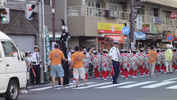 a line of people in festival dress walk down the street while a costumed person on stilts waves to onlookers